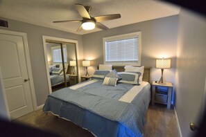 A bedroom with a bed, two bedside tables with lamps, a ceiling fan, a window with blinds, and a door.