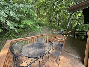 A wooden deck with a metal table and chairs, surrounded by dense greenery.