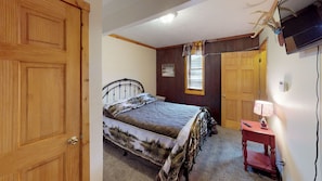 A bedroom with a bed, a wooden door, a window with blinds, a television, and a small bedside table with a lamp.