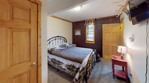 A bedroom with a bed, a wooden door, a window with blinds, a television, and a small bedside table with a lamp. - Sandy Run Cabin-Hot Tub, Hocking Hills, WNF, more (Hocking Hills)