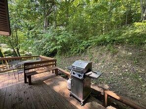 A deck with a grill, wooden bench, and metal chairs.
