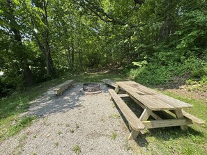 A picnic area with a fire pit, wooden benches, and picnic tables surrounded by trees.