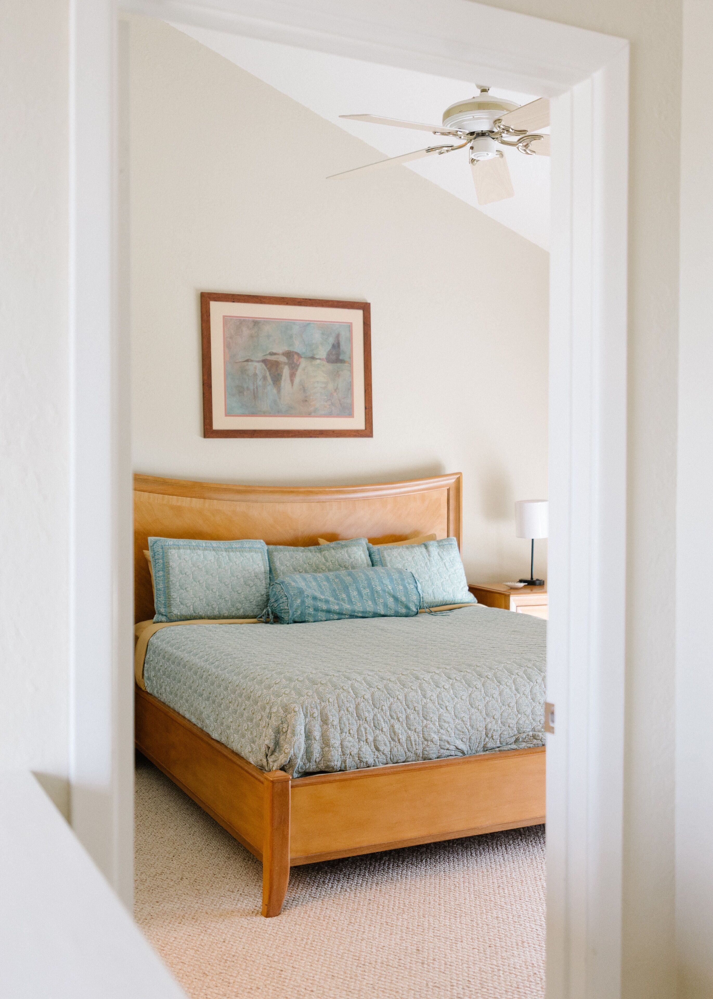 Un dormitorio con cama de madera, ventilador de techo, una obra de arte enmarcada y una mesita de noche con lámpara.