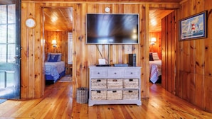 A wooden cabin interior with a TV on a dresser, two beds, and a painting on the wall.