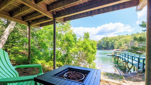 A covered outdoor seating area with a table and chairs, overlooking a river and a wooden bridge.