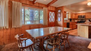 A wooden interior with a large dining table, wooden chairs, a kitchen area with wooden cabinets and appliances, and a window with curtains. - (99) Yosemite Mountain Lodge at The Redwoods In Yosemite (Wawona)