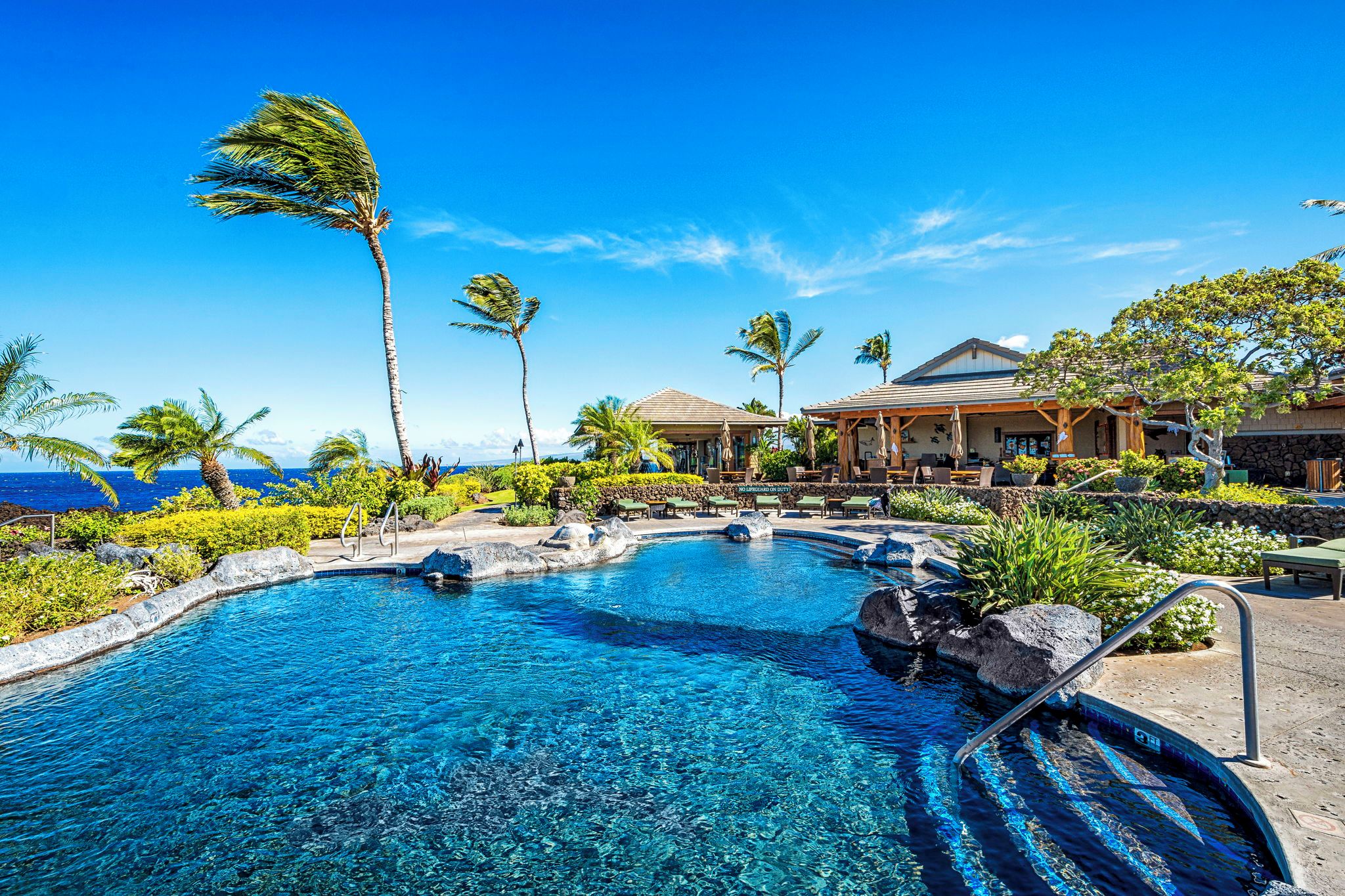 A clear blue pool with a tropical setting, surrounded by palm trees and a building with a tiled roof.