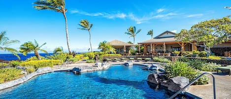 A clear blue pool with a tropical setting, surrounded by palm trees and a building with a tiled roof.