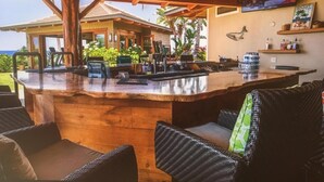 Eine Bar mit Holzbalken, Sitzgelegenheiten im Freien, einem an der Wand befestigten Fernseher und Blick auf ein Haus und Palmen.