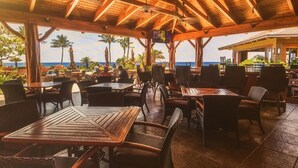 Pátio externo com mesas e cadeiras de madeira, com vista para uma praia com palmeiras e o oceano.