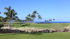 Um campo de golfe com vista clara para o oceano, palmeiras e um veículo ao longe.