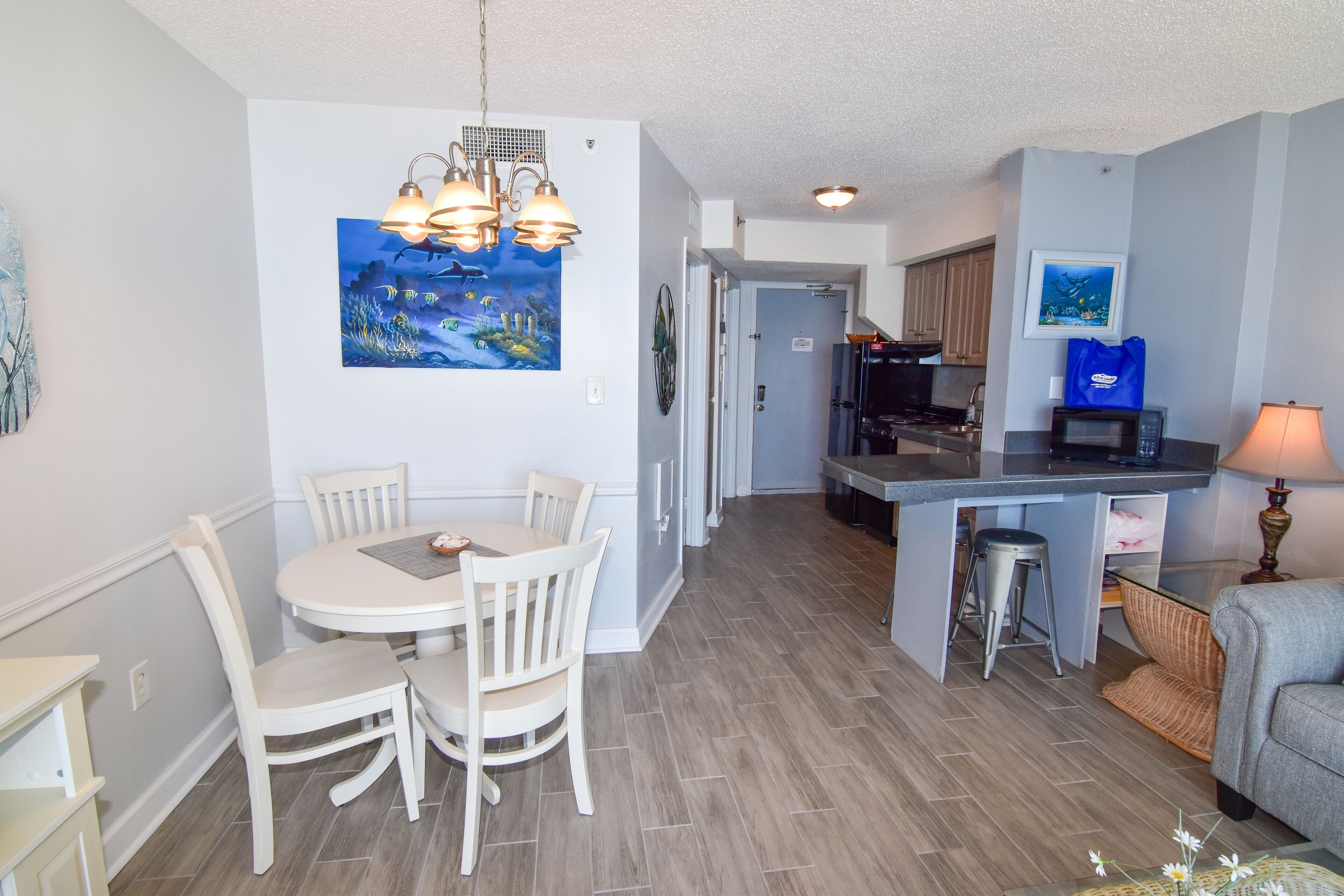 A modern kitchen with a dining area, featuring a central island, pendant lighting, and a painting on the wall.