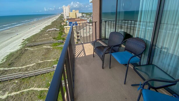 Ein Balkon mit zwei Stühlen und einem Tisch, mit Blick auf einen Strand und Gebäude in der Ferne.