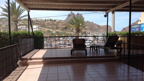 Une terrasse couverte avec un sol carrelé, deux fauteuils, une petite table et une vue sur un paysage montagneux, avec des bâtiments en arrière-plan.