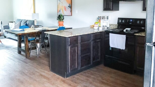 A kitchen with dark wooden cabinets, a granite countertop, a stove, and a dining area with a table and chairs. - “Ca C’est Bon” (Lake Charles)