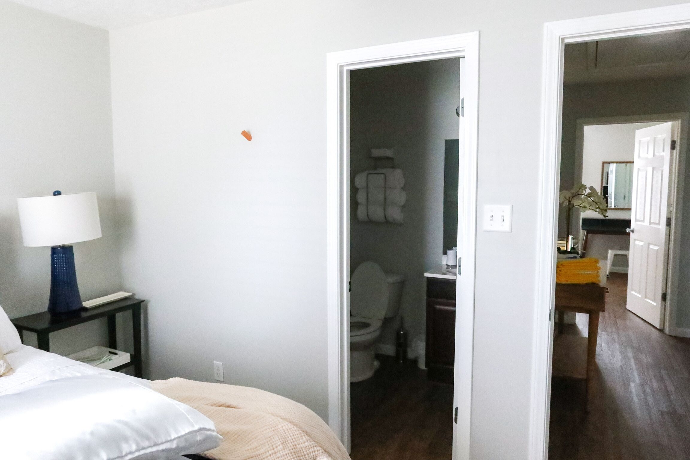 A hotel room with a bed, a nightstand with a lamp, a bathroom visible through an open door, and a hallway leading to another room.