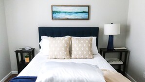 A neatly made bed with a white and beige patterned duvet, a dark blue headboard, and a framed painting above it. There is a bedside table with a lamp and a phone on it. - “Ca C’est Bon” (Lake Charles)