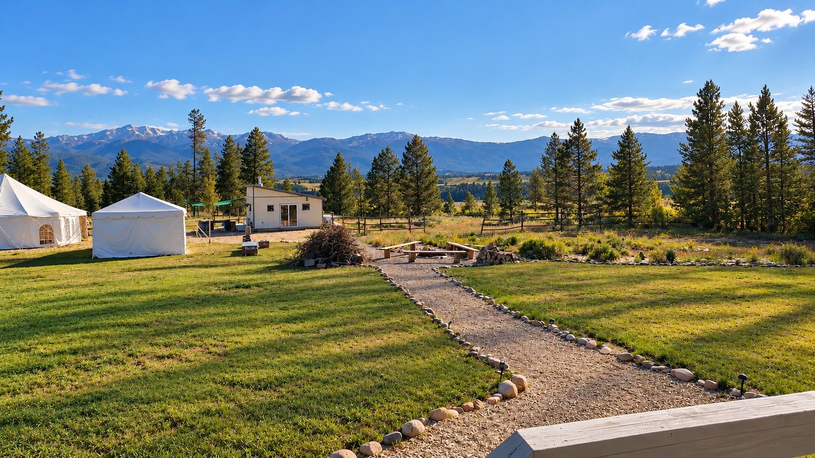 A grassy area with a paved path, white tents, and a small building, with mountains in the background.