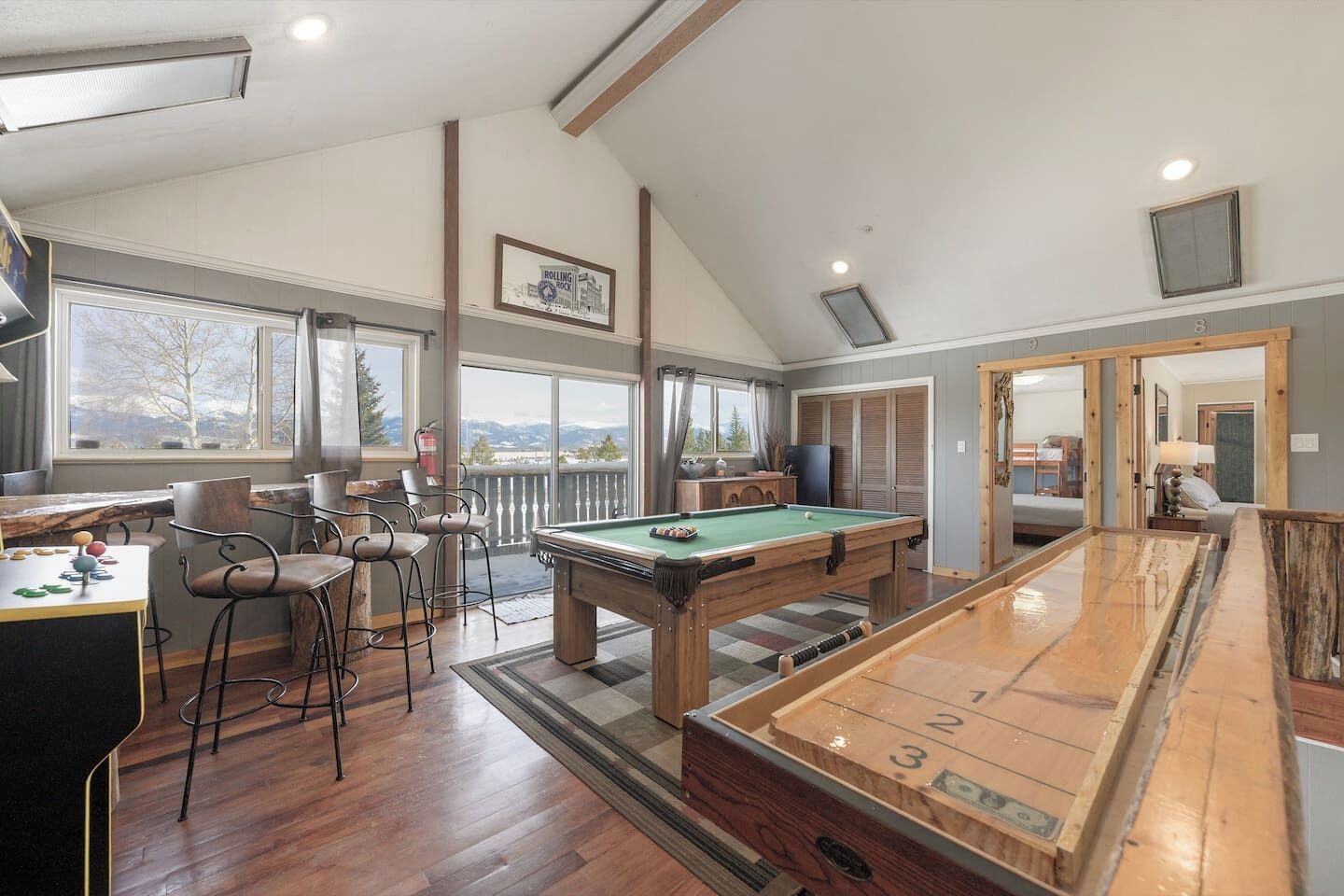 A pool table with a green felt surface, a bar area with stools, and a view through large windows.
