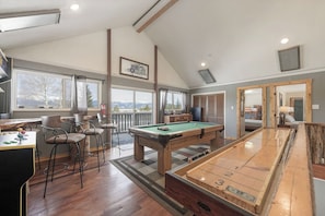 A pool table with a green felt surface, a bar area with stools, and a view through large windows. - Rustic Mountain Estate for Weddings & Retreats (Fraser)