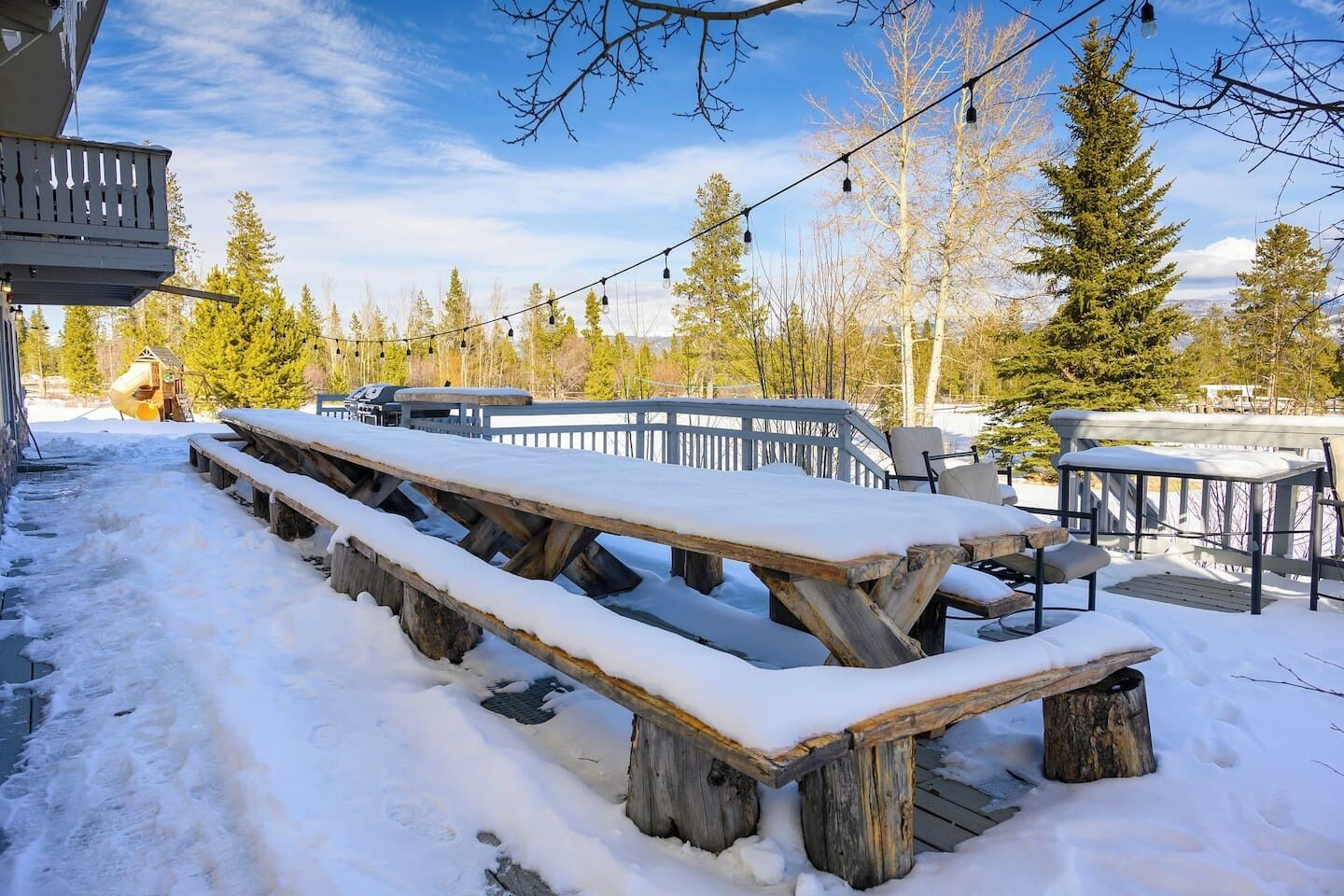 Un espace extérieur enneigé comprenant des tables et des chaises de pique-nique, une terrasse en bois et une guirlande lumineuse.
