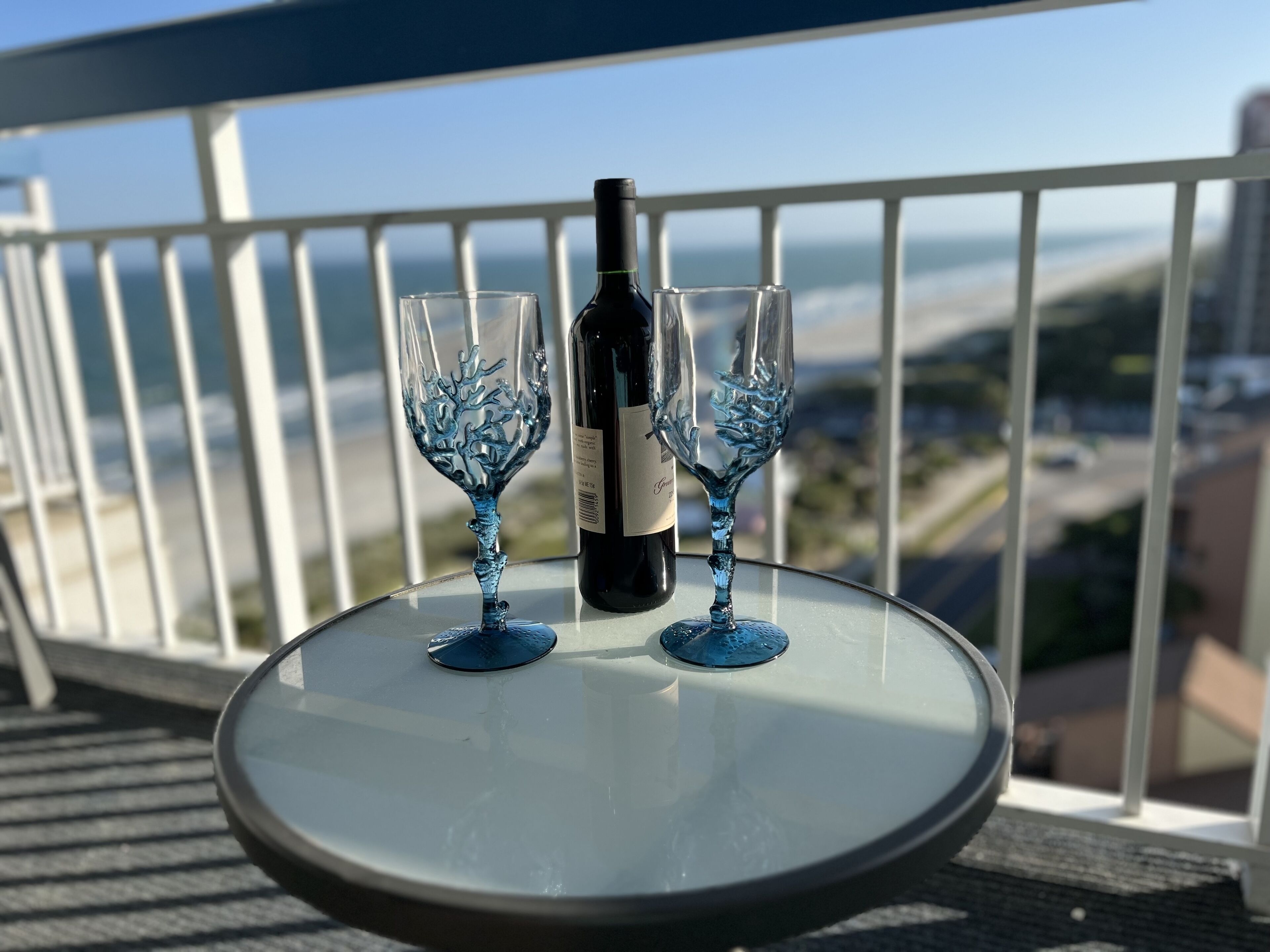 Et bord med to vinglas og en flaske på en balkon med udsigt over kysten.