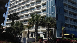 A high-rise hotel with multiple balconies, a modern glass facade, and a landscaped courtyard with palm trees and flowers. - GORGEOUS View of Ocean, Coast & Sunset! - Remodeled Forest Dunes (Myrtle Beach)