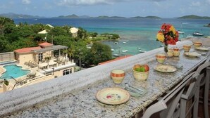 A balcony with a view of a pool, a dining table set with plates and a vase of flowers, and a coastal landscape with boats and greenery. - Grande Bay Luxury 3BR Spectacular Unobstructed View   (Cruz Bay)