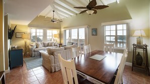 A spacious living area with a wooden dining table, white chairs, and a ceiling fan. There are large windows providing natural light and a view of the outdoors. - Grande Bay Luxury 3BR Spectacular Unobstructed View   (Cruz Bay)