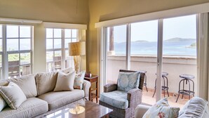 A spacious room with a large window offering a view of the sea, a comfortable sofa, a wicker chair, and a table with a lamp. - Grande Bay Luxury 3BR Spectacular Unobstructed View   (Cruz Bay)