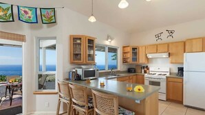 A modern kitchen with wooden cabinets, a central island with bar stools, a microwave, and a view of the ocean. - Hale Kohola ~ Spacious, Super Views - 30 Day Minim (Waimea)