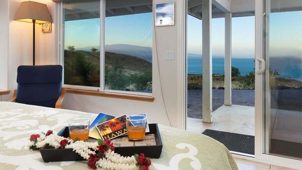 A hotel room with a bed, a desk, a chair, a lamp, a tray with flowers and a drink, and a scenic view through large windows. - Hale Kohola ~ Spacious, Super Views - 30 Day Minim (Waimea)