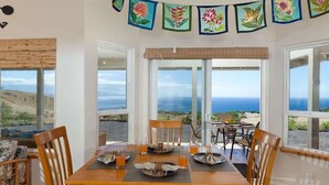 A dining area with a table set for four, large windows offering a view of the ocean, and a decorative banner with floral designs. - Hale Kohola ~ Spacious, Super Views - 30 Day Minim (Waimea)