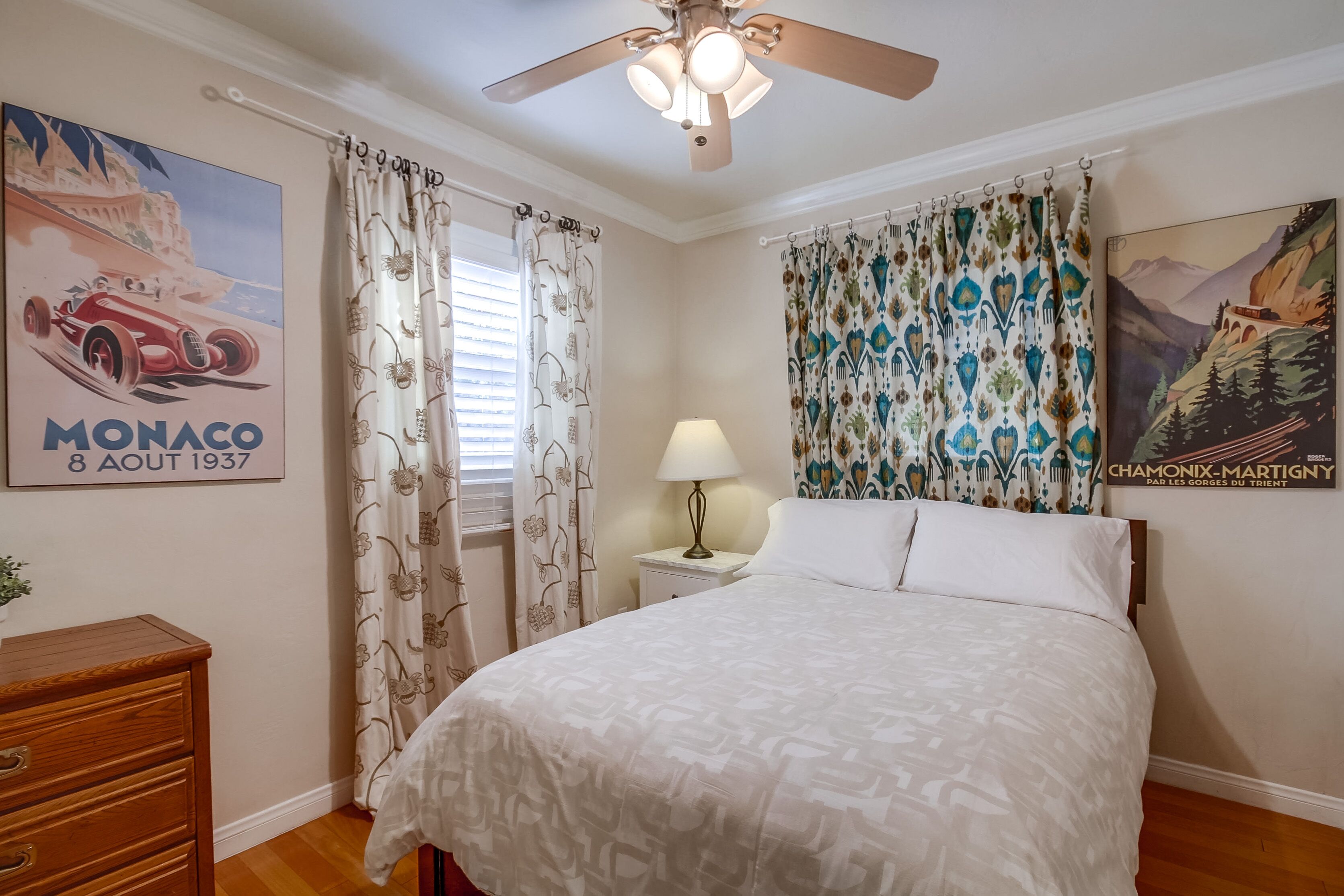 A bedroom with a bed, a nightstand with a lamp, a ceiling fan, and two curtains.
