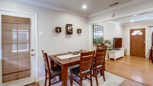 Un coin repas avec une table et des chaises en bois, un petit meuble blanc sur lequel est posée une télévision, et une porte dotée d’une moustiquaire.