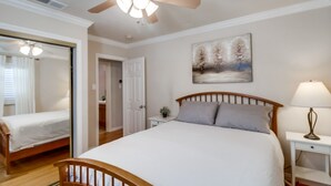 Une chambre à coucher comprenant un lit, une table de chevet, une lampe, un ventilateur de plafond, une porte et un tableau accroché au mur.