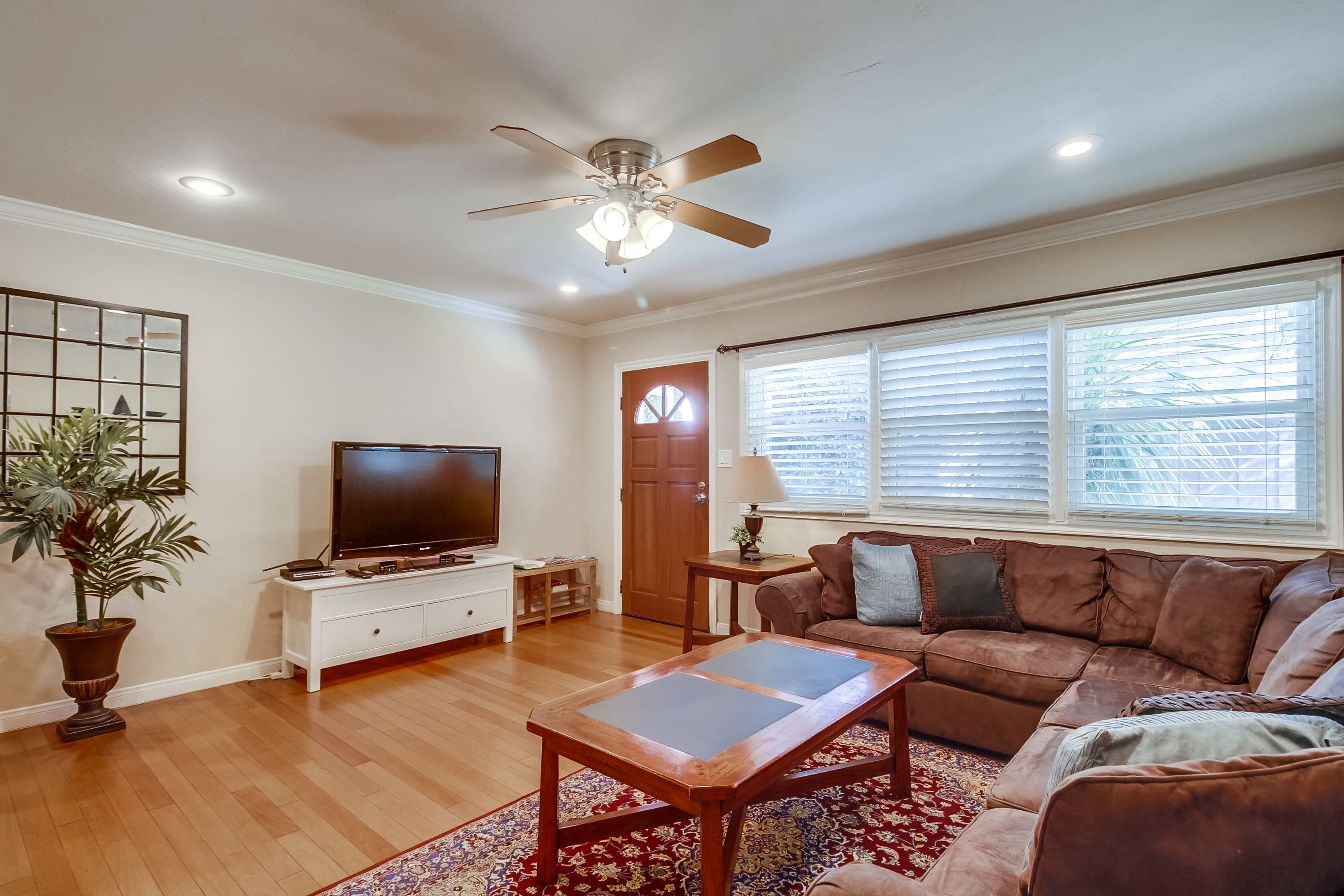 A living room with a ceiling fan, a TV on a white cabinet, a brown leather couch, and a red patterned rug.
