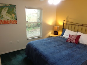 A bedroom with a bed, a nightstand, a lamp, a window, and a wall hanging.
