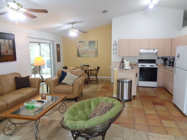 A living room with a sofa, armchair, and a glass coffee table. There is a dining area with a table and chairs. The kitchen has a stove, sink, and cabinets.