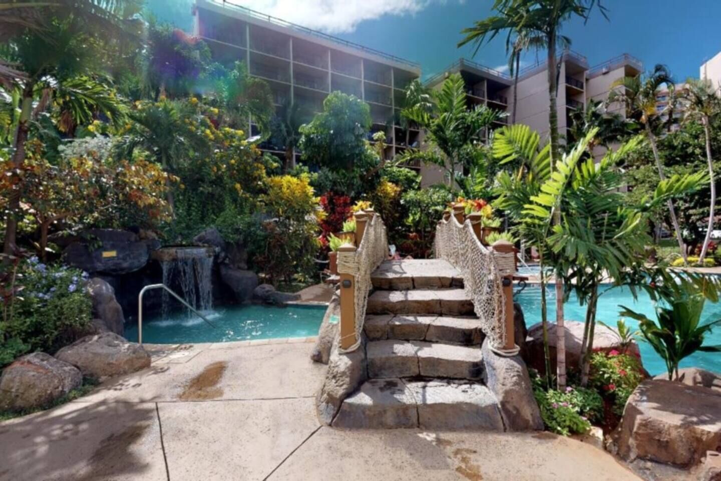 Uma área de piscina com cachoeira, degraus de pedra e plantas tropicais.