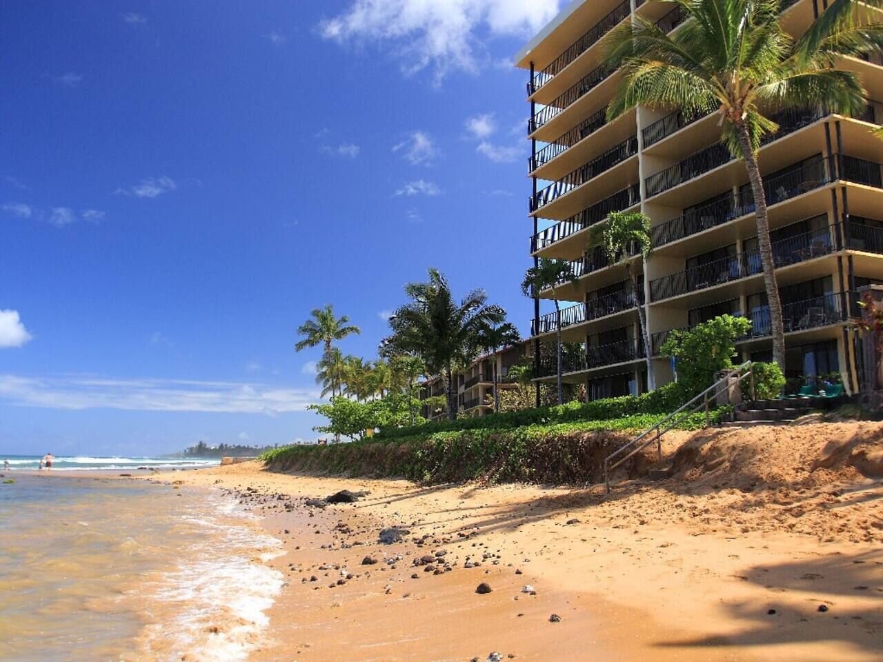 Ein Hotel direkt am Strand mit mehreren Balkonen, Palmen und einem Sandstrand.