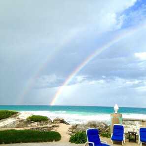 Blick auf einen Strand mit Regenbogen, blauen Liegestühlen und einem Steinpfeiler.