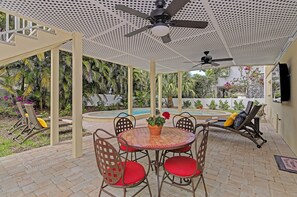 En overdækket terrasse med et bord og stole, en loftsvifte samt udsigt til en swimmingpool og grøn bevoksning.