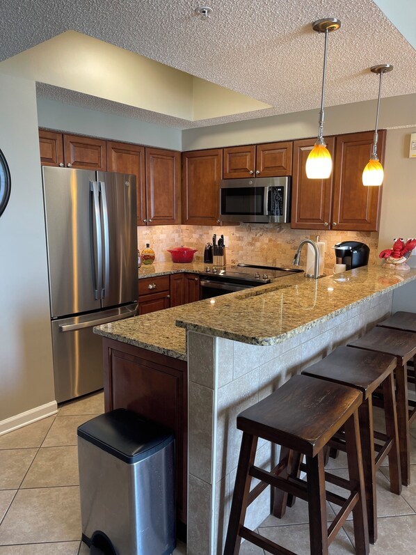 A modern kitchen with granite countertops, stainless steel appliances, wooden cabinets, and bar stools.