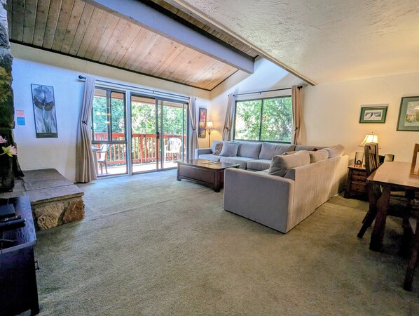 Ein gemütliches Wohnzimmer mit Sofa, Couchtisch und Esstisch. Große Fenster mit Vorhängen, ein Steinkamin und Blick ins Freie.