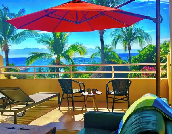 A balcony with a red umbrella, two chairs, a small table, and a view of the ocean and palm trees.