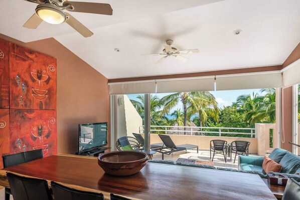 A modern living room with a wooden dining table, a large bowl, and a TV. There are two ceiling fans, a balcony with a view of palm trees, and a sofa with cushions.