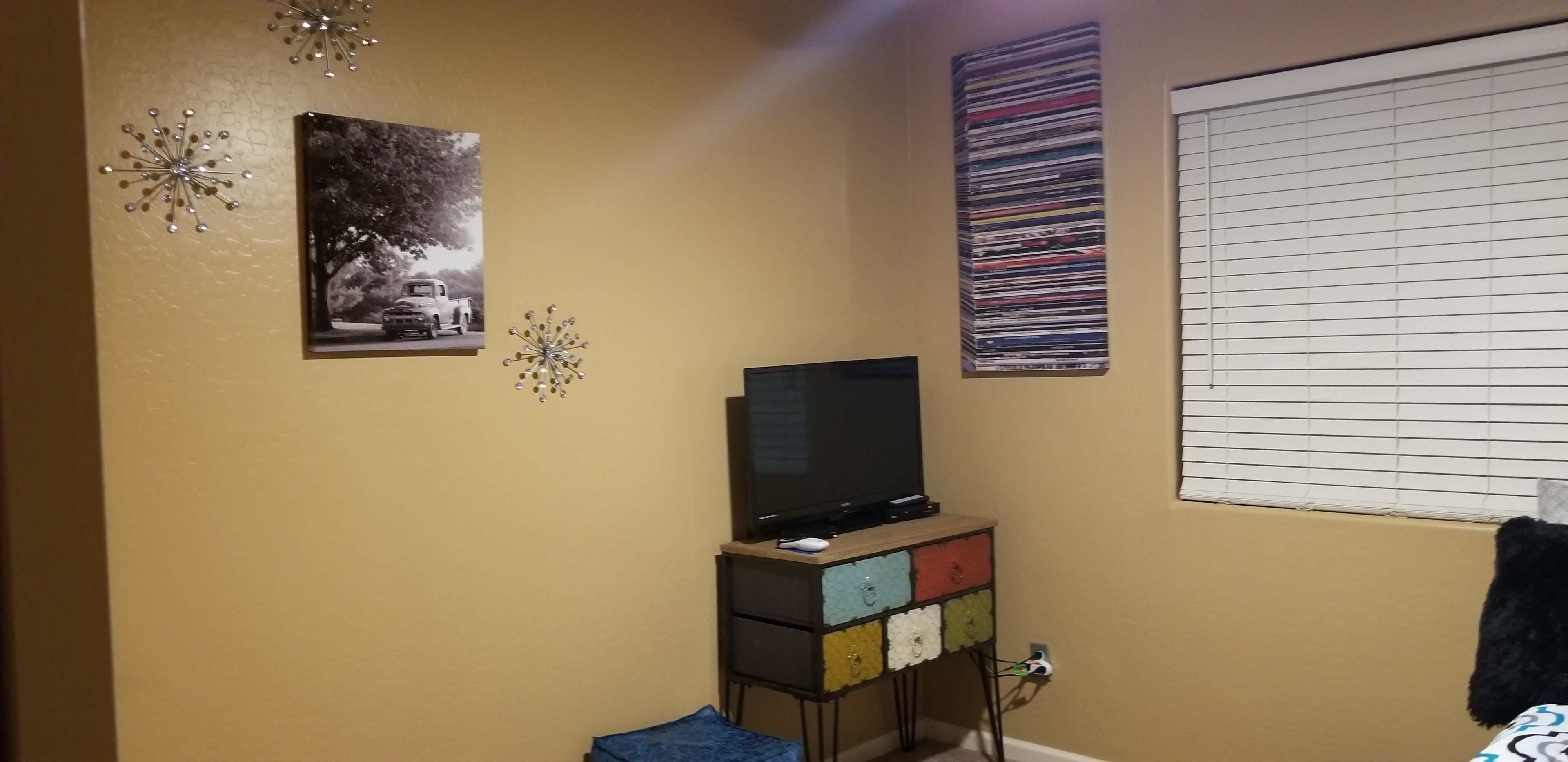 A room with a television on a wooden stand, a wall-mounted shelf with colorful drawers, a window with blinds, and two wall decorations.
