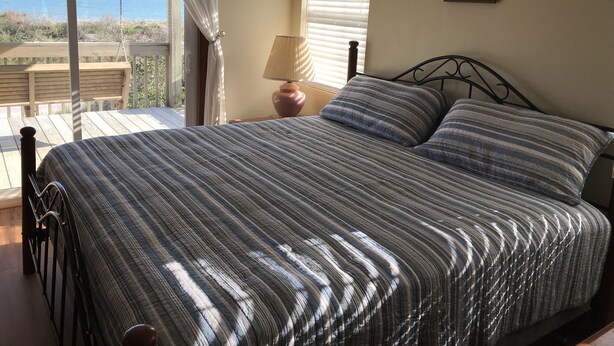 A bedroom with a bed, a nightstand, a lamp, and a view of the sea. - Mother Ocean, Paradise, Oceanfront Panoramic Ocean Views, Private Beach (Holly Ridge)