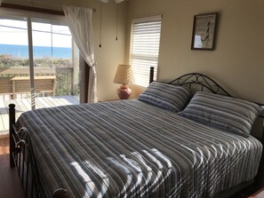 A bedroom with a bed, a nightstand, a lamp, and a view of the sea. - Mother Ocean, Paradise, Oceanfront Panoramic Ocean Views, Private Beach (Holly Ridge)